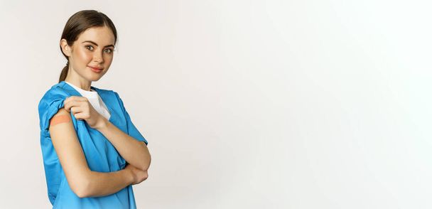 Female nurse, doctor medical worker got vaccinated, showing shoulder with patch after covid-19 vaccine, smiling pleased, standing over white background. - Photo, image