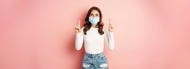 Portrait of young glamour girl in medical face mask, reading promo text, pointing and looking up, standing over pink background. Covid and pandemic concept - Photo, image