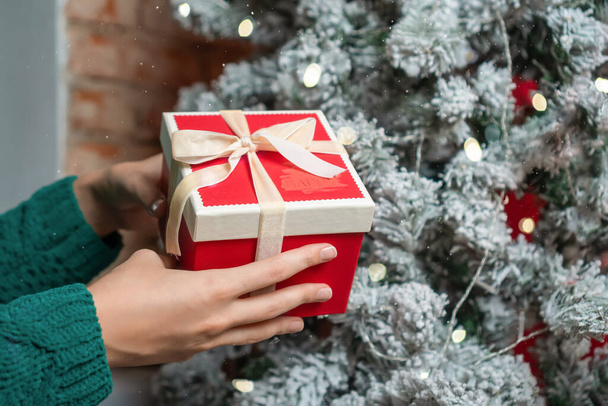 Las manos de las mujeres en un suéter verde sostienen una caja de regalo de Navidad roja con un lazo cerca de un árbol de Navidad. Concepto de Navidad y Feliz Año Nuevo - Foto, imagen