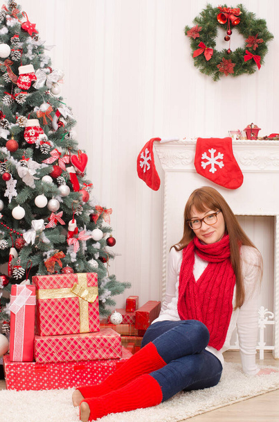 白いセーターを着た長髪の少女と赤いスカーフとゴルフは、明るい部屋の暖炉の背景を背景に、クリスマスツリーとギフト付きの箱の近くに座っています。 - 写真・画像