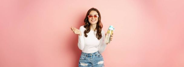 Stylish girl in spring outfit, wearing sunglasses, holding water bottle with lemon, healthy drink, laughing and smiling, standing over pink background. - Photo, Image