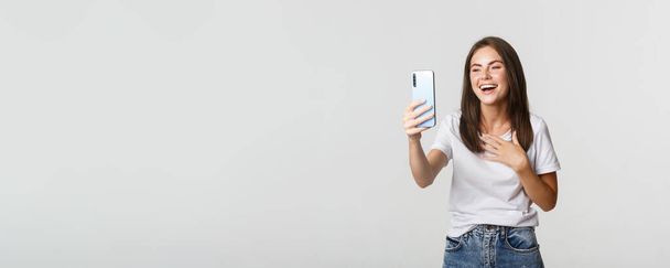 Friendly smiling woman video calling and talking on smartphone happy, standing white background. - Photo, Image