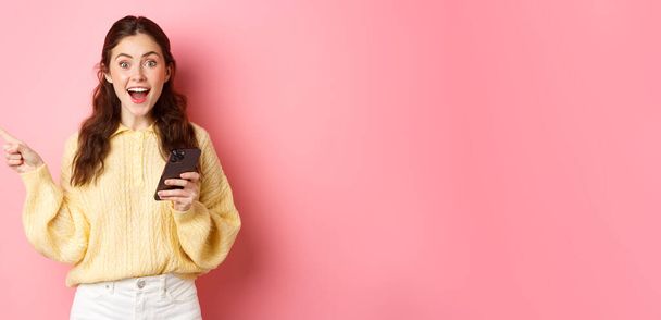 Excited young woman holding mobile phone, pointing finger aside at left copyspace, showing advertisement, found interesting promo online in smartphone, standing against pink background. - Foto, Bild