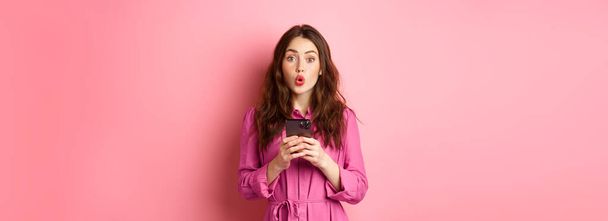 驚きの若い女性が息をのむとすごいと言ってのイメージは、カメラでピンクの背景にスマートフォンで立って、携帯電話上のニュースをオンラインで読んだ後に感銘を見つめている。スペースのコピー - 写真・画像