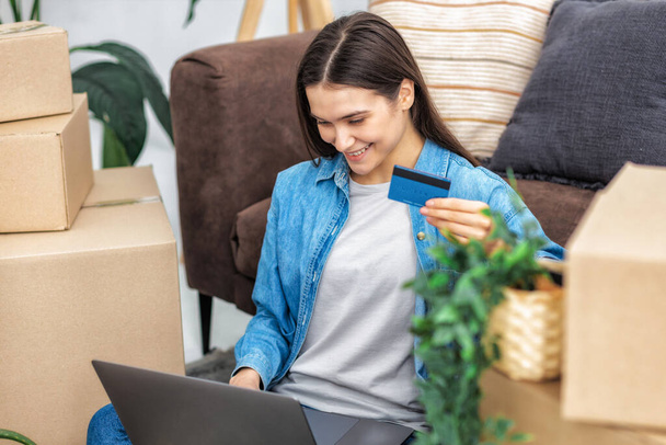 Moving, new house concept. Happy young caucasian woman sitting on the floor with laptop and credit card orders furniture online in her new home - Foto, afbeelding