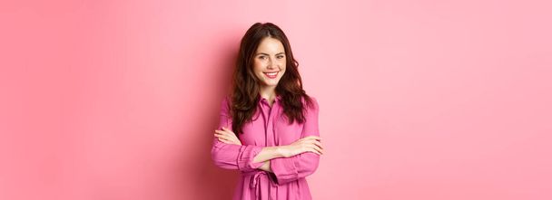 Gyönyörű nőies nő tavaszi ruha, kézen fogva a mellkason, és mosolyogva magabiztos kifejezés, álló elégedett rózsaszín háttér. - Fotó, kép