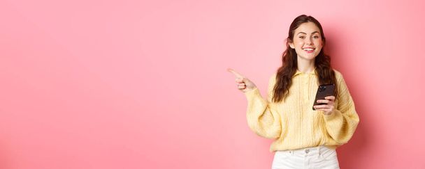 Beautiful modern girl holding smartphone and pointing finger aside, showing promotional text on copyspace while using mobile phone, standing over pink background. - Fotografie, Obrázek