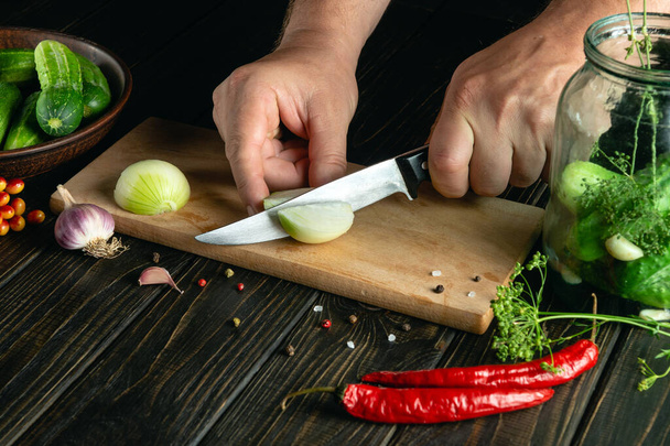 Ο μάγειρας κόβει κρεμμύδια σε ένα ξύλο κοπής για τουρσί σε ένα βάζο με αγγούρια και μπαχαρικά. Μενού για ένα εστιατόριο ή ξενοδοχείο σε σκούρο φόντο - Φωτογραφία, εικόνα