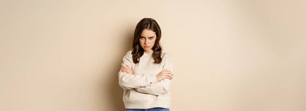 Angry girlfriend sulking, cross arms on chest and look offended, staring moody at camera, standing over beige background. - Foto, afbeelding