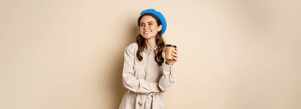Kaunis moderni tyttö poseeraa noutoruokaa kuppi kahvia, juo uudelleenkäytettäviä kuppi kahvilasta ja poseeraa onnellinen, beige studio tausta. - Valokuva, kuva