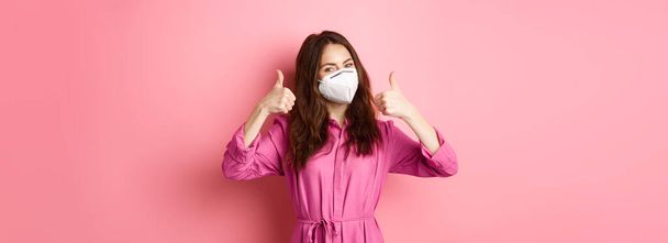 Covid-19, pandemic and lifestyle concept. Cheerful girl shows thumbs up, wears medical respirator as preventive measure from corona, pink background. - Foto, imagen