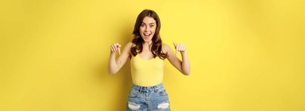 Look down, big news. Portrait of amazed young woman pointing down below, smiling excited, showing logo or banner, standing over yellow background. - Foto, afbeelding