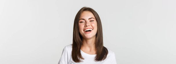 close-up van gelukkig brunette meisje in wit t-shirt lachen en glimlachen zorgeloos op camera. - Foto, afbeelding