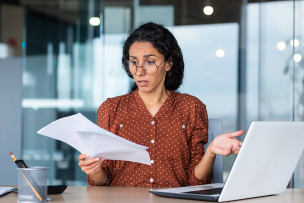 Frustrated and sad business woman holding financial report in hands, Hispanic woman sad working in office using laptop, female financier accountant unhappy with work results. - Foto, immagini