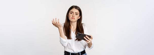 Sad woman losing in mobile video game, looking upset and disappointed at smartphone, sulking, standing over white background. - Фото, изображение