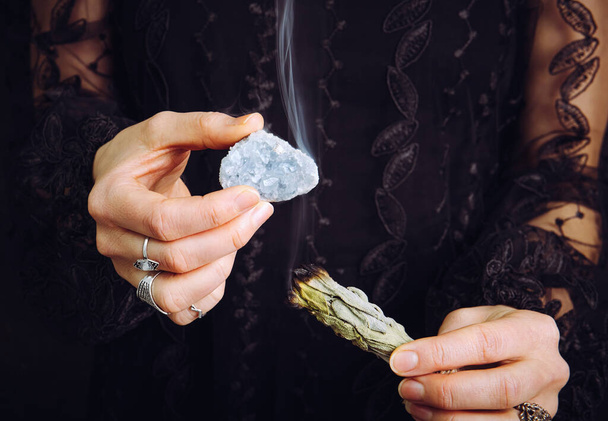 Close up view of woman in black lace dress, cleansing blue celestite  crystal cluster gemstone by smudging white sage bundle. Remove negative energy, cleanse crystal. - Fotoğraf, Görsel