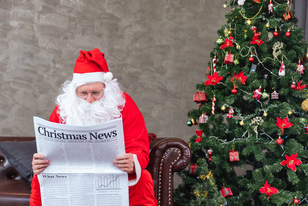 Santa Claus reading Christmas news from the newspaper while sitting on the sofa couch by the christmas tree for long holiday economics situation and accident traffic transport report - Photo, Image