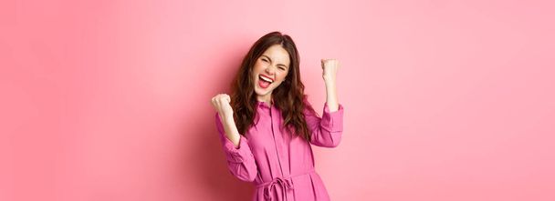 Beautiful smiling woman scream with happy and excited face, saying yes, making fist pump, winning and feeling like champion, triumphing, standing over pink background. - Photo, Image