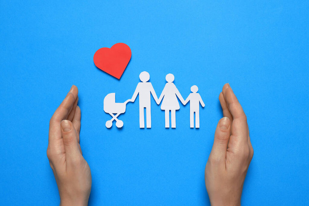 Woman protecting paper family cutout on light blue background, top view. Insurance concept - 写真・画像