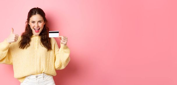 Jonge moderne vrouw die ja zegt en duimen laat zien, plastic kredietkaart vasthoudt, over een roze achtergrond staat. Kopieerruimte - Foto, afbeelding