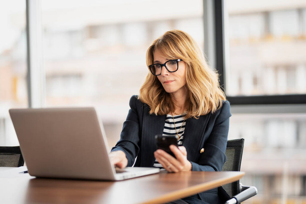 Confident businesswoman using mobile phone and laptop at the office. Attractive blond haired professional female wearing glasses and blazer. - Foto, afbeelding