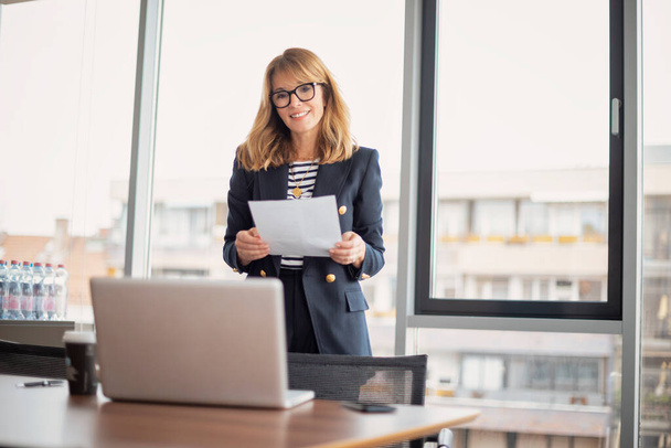 Shot of a confident mature businesswoman having video call while standing in a modern office and holding document in her hand. Professional female wearing eyeglasses and blazer. - Valokuva, kuva