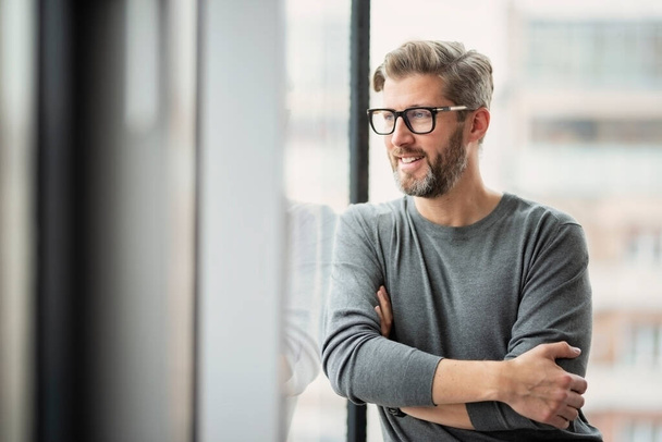 Portrait of confident businessman wearing glasses and sweater while standing at the office and looking out the window.  - Valokuva, kuva