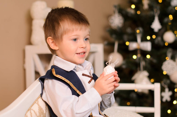 Gülümseyen çocuk kanepede oturuyor. Beyaz bir gömlek, siyah bir yelek giyip Noel ağacının arka planında şen şakrak giyinmiş. - Fotoğraf, Görsel