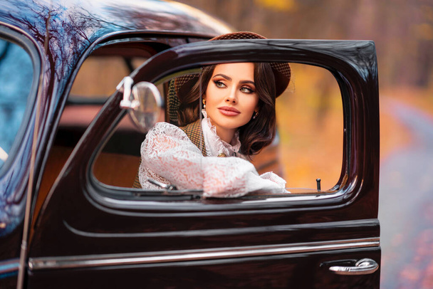 Beautiful female in vintage  dress, lace blouse and hat with veil sitting in retro brown car on autumn background. Elegant lady in gloves posing on nature.Warm art work. - Foto, Imagen