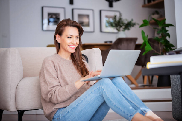 Full length of an attractive middle aged woman sitting on floor while using laptop. Beautiful female wearing casual clothes.  - Фото, зображення