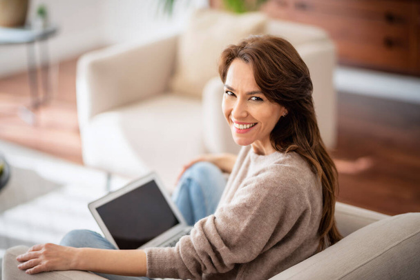 Close-up of an attractive middle aged woman sitting on couch while using laptop. Beautiful female wearing casual clothes.  - Photo, Image