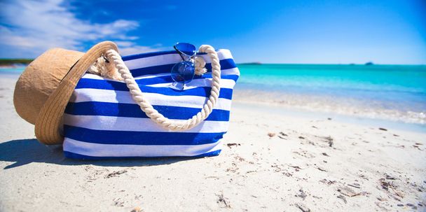 Смугастий мішок, солом'яний капелюх, сонцезахисний крем і рушник на білому тропічному пляжі
 - Фото, зображення