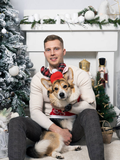 A young man with a corgi dog is sitting by a Christmas tree against the background of a fireplace. A dog in a Christmas costume. Happy holiday time. - Photo, Image