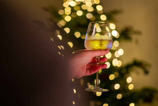 Ręczne trzymanie kieliszka wina, rozmazane lampki świąteczne na choince w tle. Sosna ozdobiona świecącą girlandą, osoba trzymająca kubek z napojem. - Zdjęcie, obraz