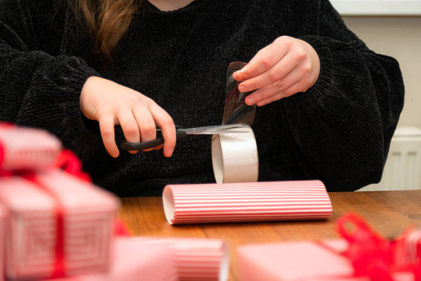 Kobieta pakująca prezent w papier ze striptizem z czerwoną wstążką. Ręce pakujące pudełko w czerwony i biały papier świąteczny. - Zdjęcie, obraz
