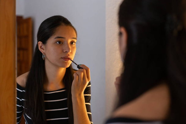 Joven señora étnica mirando en el espejo mientras se aplica brillo labial  - Foto, imagen