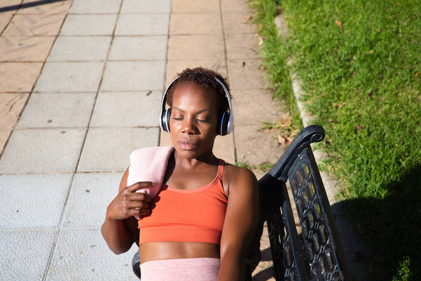 Jovem afro-americana sentada no banco descansando depois de praticar esporte no parque com toalha sobre o ombro e ouvindo música com fones de ouvido brancos. Conceito de esporte e saúde. - Foto, Imagem