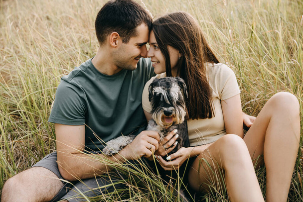 Szczęśliwa para mężczyzn i kobiet siedzących na zewnątrz, przytulających miniaturowego psa czystej krwi sznaucera, uśmiechających się. - Zdjęcie, obraz