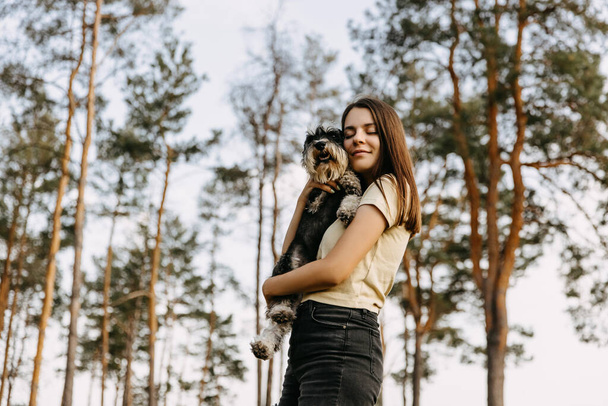 Νεαρή μελαχρινή γυναίκα αγκαλιάζει ένα μικροσκοπικό σκυλί ράτσα Schnauzer, χαμογελώντας. - Φωτογραφία, εικόνα