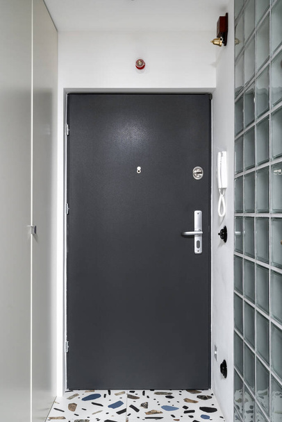Vertical shot of black metal door on white wall with intercom communication system, glass brick elements, wardrobe for outerwear in corridor.Hallway with minimalist interior design in modern apartment - Photo, Image