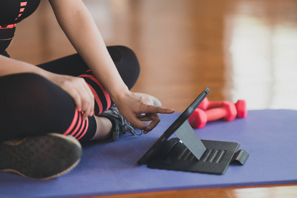 Gewasfoto. Handsporter aanraken van tablet zitten op yoga mat en doen oefening met halter workout online en kijken video streaming op tablet in de fitnessruimte. Online fitness concept. - Foto, afbeelding