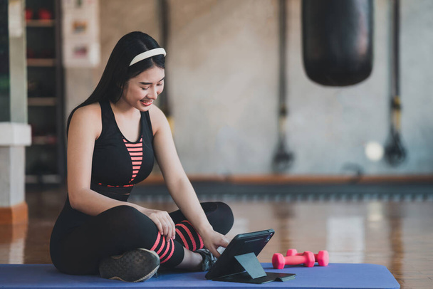Молодая азиатская спортсменка сидит на коврике для йоги и делает упражнения с гантели тренировки онлайн и глядя видео потокового на планшете в помещении фитнес-тренажерный зал. Онлайн тренировки по фитнесу. - Фото, изображение