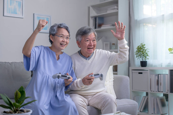 Happy Asian senior couple sitting together on their living room sofa holding controllers and laughing while playing a video game. - Photo, Image