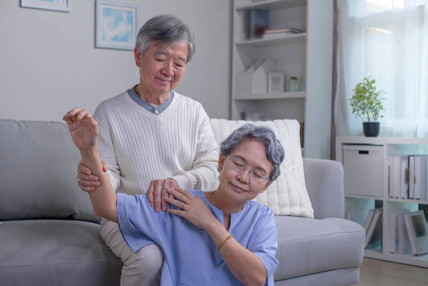 Asiática viejo senior anciano esposa sentado en piso teniendo problema con sufrir dolor de espalda doloroso hombro mientras cuidado marido de pie detrás ayuda masaje espalda. - Foto, imagen