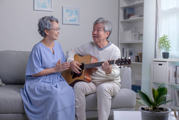 自宅でギターを弾いている間に、アジアの成熟した男性と女性が好きな歌を歌う。自宅のリビングルームでソファに座っている間、高齢者や女性は楽しい時間を過ごしています。引退、人生、ライフスタイル、ケアフリー. - 写真・画像