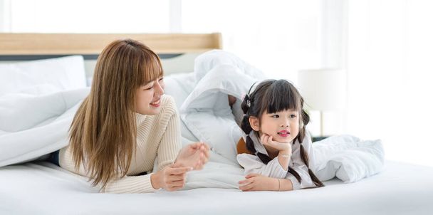 Millennial Asian young beautiful mother in sweater waking up laying down smiling together under thick warm blanket on bed in morning with little cute preschooler daughter girl in bedroom at home. - Valokuva, kuva