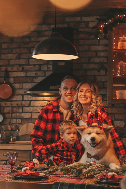 Portrait of happy family with son in red plaid pajamas laughing hugs kisses at home eve 25 december. Celebrating new year noel cozy snuggle warm weather garlands lights hugging his cute akita inu dog - Photo, Image