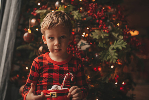Happy little boy dreamer waiting at miracle Santa enjoy sweets color lollipop. Red checkered sleepwear cheerful kid celebrating new year hold Xmas cup with marshmallows near noel tree eve 25 december - Foto, Imagen