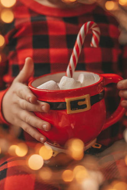 Onnellinen pieni poika uneksija odottaa ihme Joulupukki nauttia makeisia väri tikkari. Punainen ruudullinen yöpuvut iloinen lapsi juhlii uutta vuotta pidä Xmas cup vaahtokarkkeja lähellä noel tree eve 25 joulukuu - Valokuva, kuva