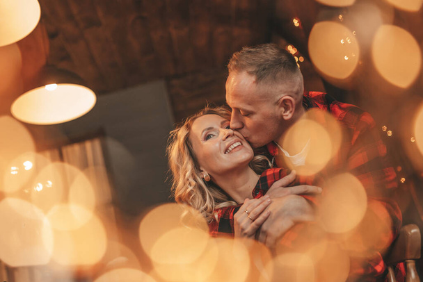 Portrait of young lovely couple hugging indoor eve 25 December. Lovers laughing hugs kisses waiting miracle at home. Celebrating new year garlands lights noel in red plaid pajamas passion tenderness - Photo, Image